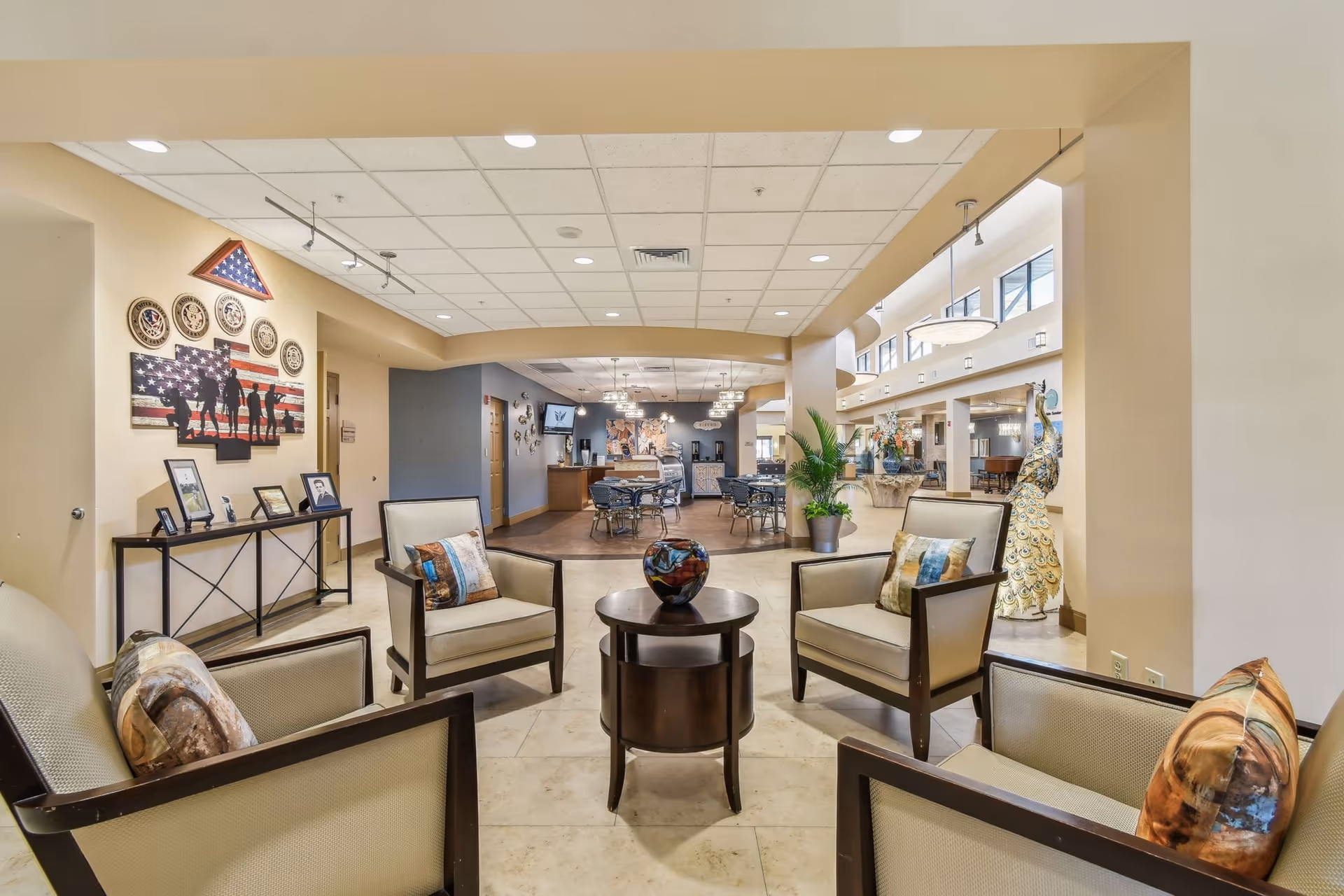 A spacious and well-lit common area in a senior living facility featuring four cushioned armchairs arranged around a small round wooden table with a decorative vase. In the background, there are dining tables and chairs, wall decorations including patriotic artwork and framed photos, and large windows letting in natural light. The ceiling has recessed lighting and hanging fixtures, and there are plants and sculptures adding to the decor.