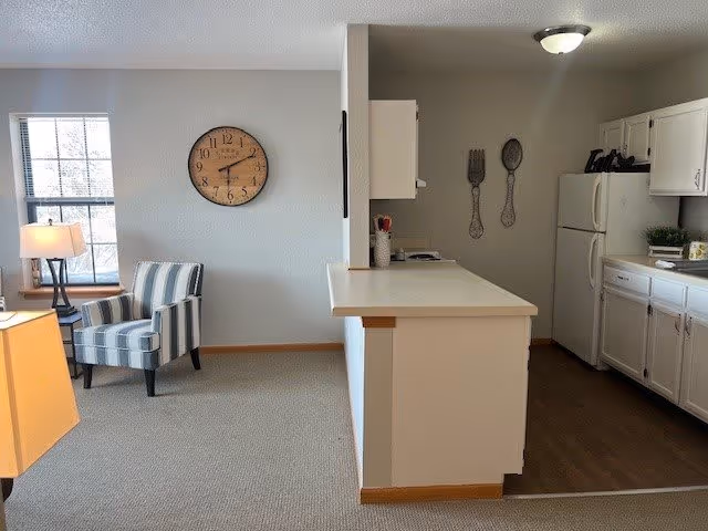 Interior view of a senior living facility showing a small living area with a striped armchair and a lamp next to a window. A large wall clock is mounted on the wall. Adjacent to the living area is a kitchen with white cabinets, a refrigerator, and a countertop bar separating the two spaces. The kitchen floor is wooden, while the living area is carpeted.