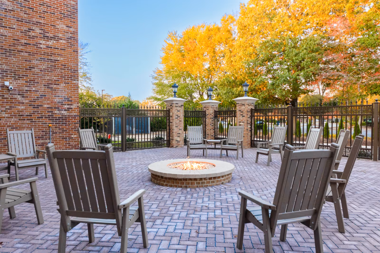 Paved outdoor patio area with a central circular fire pit surrounded by wooden chairs, brick columns, a metal fence, and trees with autumn foliage.