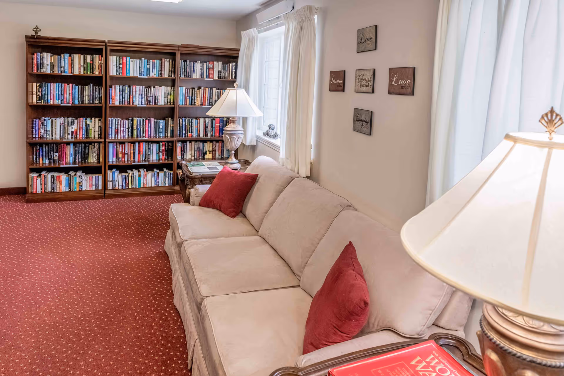 A cozy living room area with a beige couch adorned with red pillows, a wooden side table with a lamp, and a large bookshelf filled with books. The room has a red carpet and light-colored walls decorated with inspirational word plaques. A window with white curtains lets in natural light.