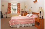 Furnished senior bedroom with a twin bed in a striped pink cover, wooden nightstands and dresser, lamps, a TV, and a window with floral curtains.