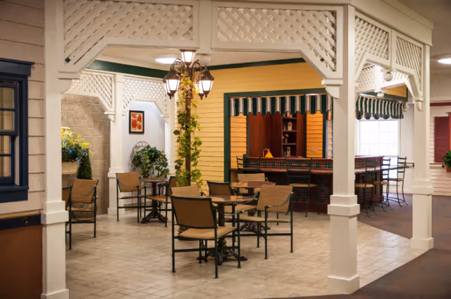 Indoor seating area with several tables and chairs arranged under a white lattice pergola structure. There is a decorative lamp post with hanging plants in the center. In the background, there is a counter with bar stools and a striped awning above it. The space has a cozy, inviting atmosphere with warm lighting and decorative plants.