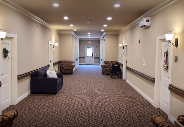 A long, well-lit hallway in an assisted living facility with beige walls and patterned carpet. The hallway features several white doors decorated with wreaths and wall-mounted lights. There are multiple seating areas with armchairs and a small couch along the walls. A wheelchair is parked near one of the doors, and an exit door is visible at the far end of the hallway.