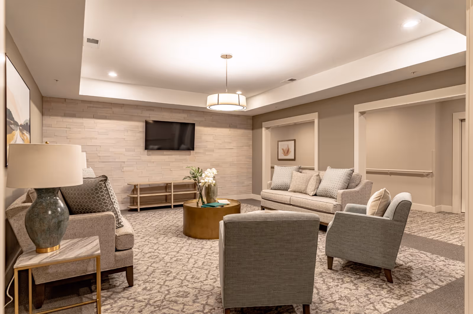 A cozy living room area with two beige sofas and two gray armchairs arranged around a round gold coffee table with a vase of flowers. The room has a patterned carpet, a wall-mounted flat-screen TV on a textured stone accent wall, a side table with a lamp, and neutral-colored walls with framed artwork.