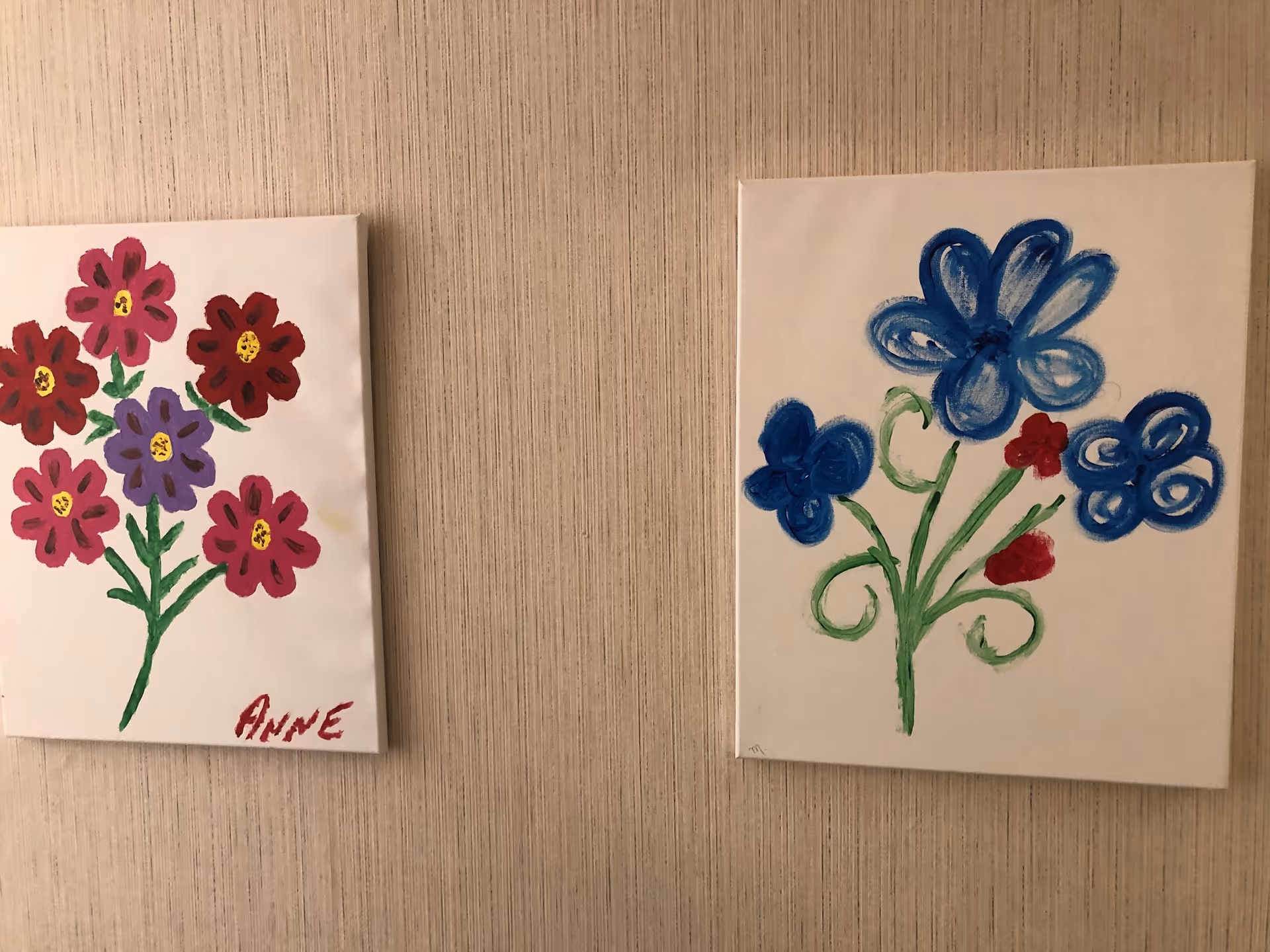 Two paintings of flowers hanging on a beige textured wall. The painting on the left features red and purple flowers with green stems and is signed 'ANNE'. The painting on the right shows blue and red flowers with green stems.