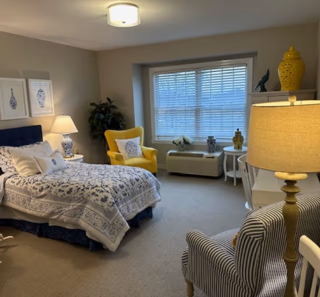 Well-lit furnished bedroom featuring a bed with patterned bedding, a yellow accent chair, a striped armchair, lamps, and a large window with blinds.