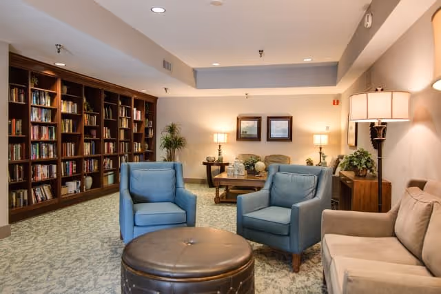 A cozy living room area with two blue armchairs, a beige sofa, and a round brown leather ottoman in the center. The room features a large wooden bookshelf filled with books along one wall, two framed pictures on the far wall, and several lamps providing warm lighting. There are also some plants and decorative items on tables around the room.