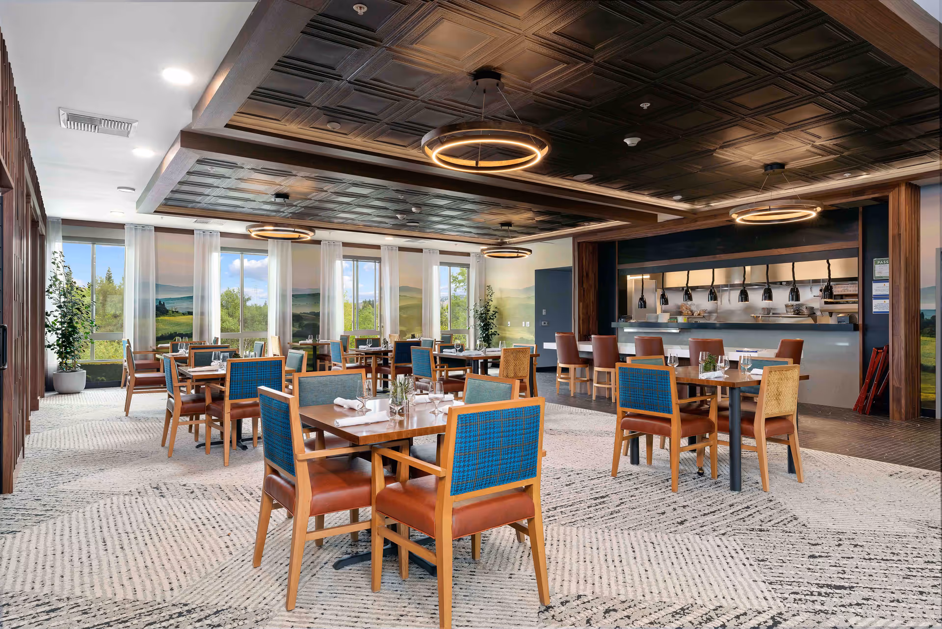A spacious dining room with multiple wooden tables and chairs featuring blue and brown upholstery. The room has large windows with sheer white curtains allowing natural light to brighten the space. The ceiling has a dark, textured design with modern circular light fixtures. In the background, there is a counter area with high chairs and hanging lights, likely for food service or a bar area. The carpet has a patterned design in neutral tones.
