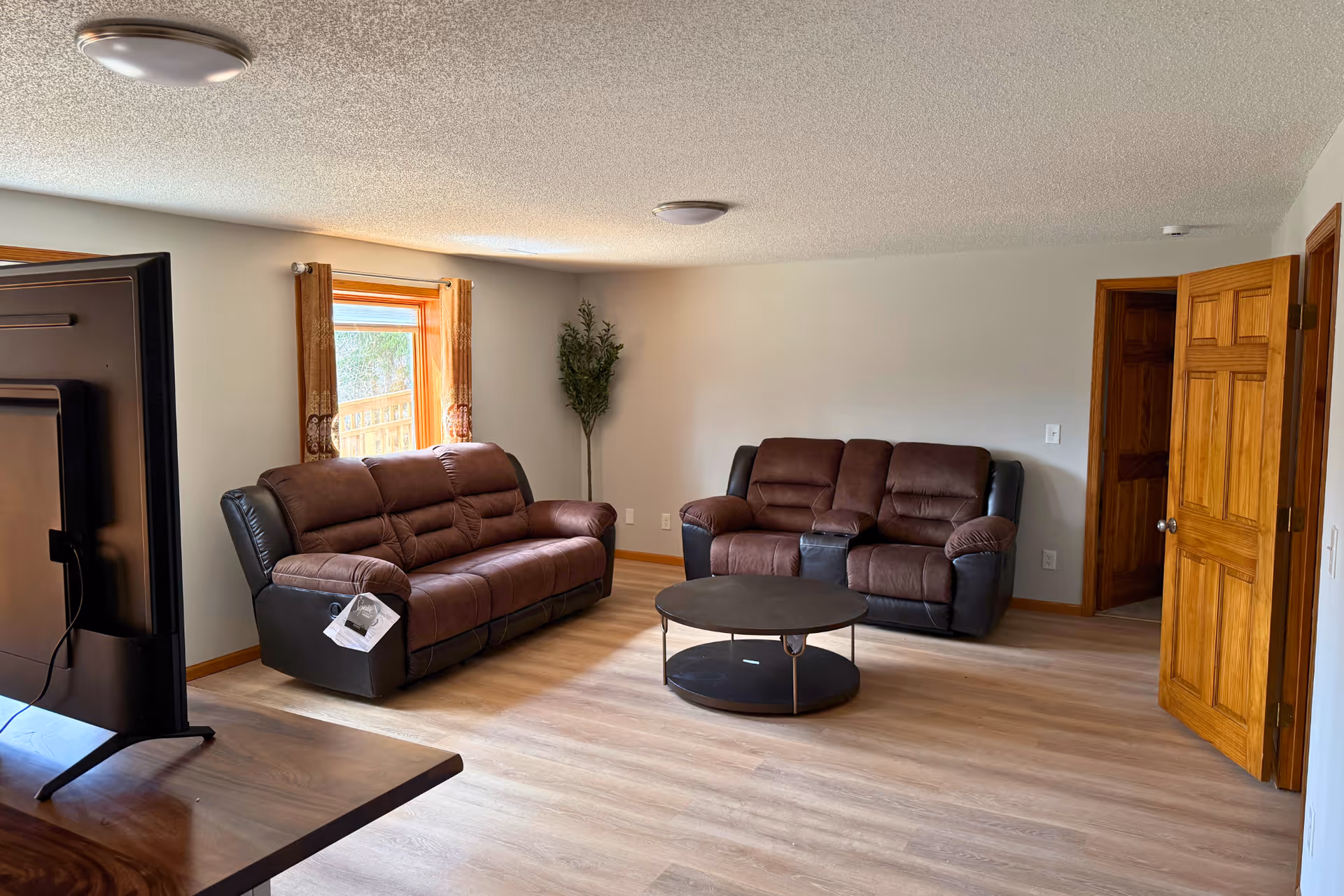 A living room with two brown leather sofas, a round black coffee table, a window with brown curtains, a potted plant in the corner, and a wooden door partially open. A flat-screen TV is visible on the left side of the image.