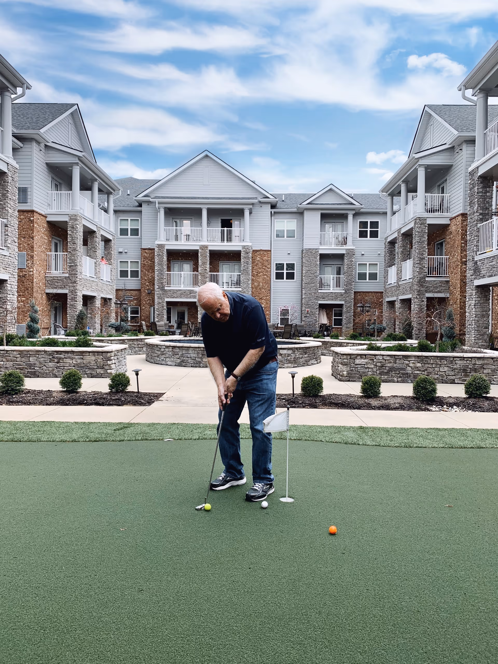 An older man putting on a small artificial putting green in the courtyard of a multi-story senior living apartment building.