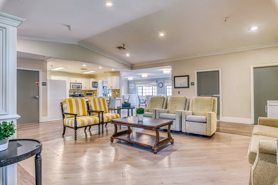 Spacious senior living common area with upholstered chairs, a coffee table, and an open view into a kitchen/dining area.