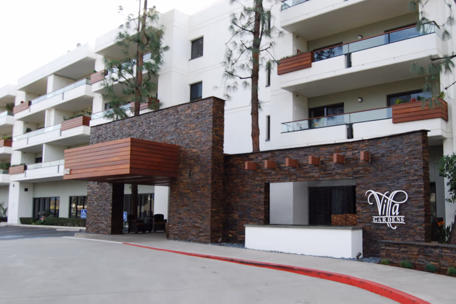 Exterior view of a modern multi-story senior living facility with white walls and balconies featuring wooden accents. The entrance has a stone facade with a wooden canopy and a sign that reads 'Villa Gardens'. There are tall trees near the building and a driveway with a red curb in front.