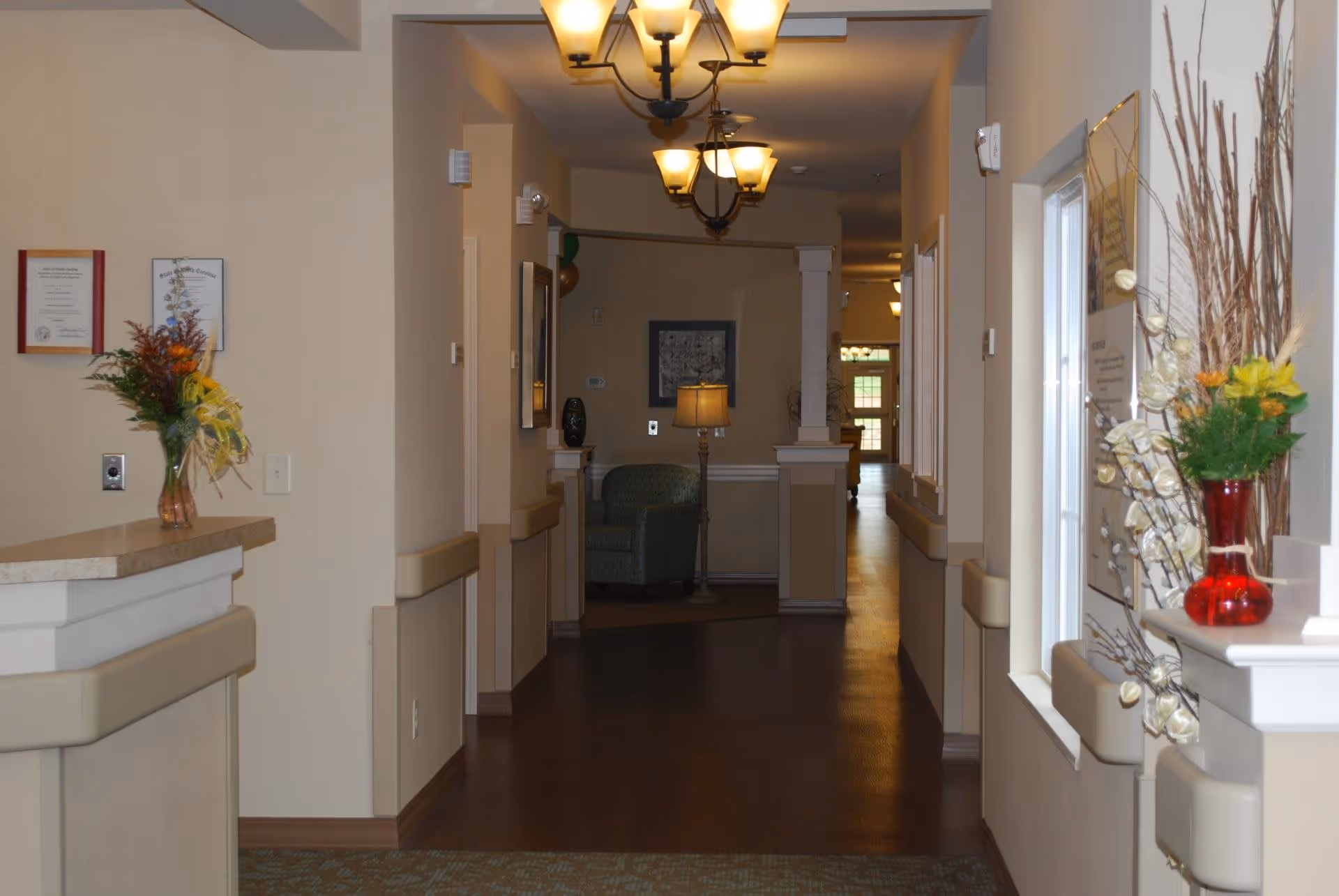 Interior hallway of a senior living facility with beige walls, wooden flooring, and soft lighting from ceiling fixtures. There are handrails along the walls, a small reception counter with a flower vase on the left, and a window with decorative plants on the right. At the end of the hallway, there is a green armchair and a floor lamp.