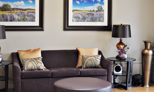 A cozy living room area with a dark brown sofa adorned with two beige and two patterned pillows. In front of the sofa is a round ottoman. On either side of the sofa are black side tables, each with a decorative lamp and a small flower arrangement. Above the sofa hang two framed landscape photographs featuring fields of purple flowers under a blue sky with clouds.