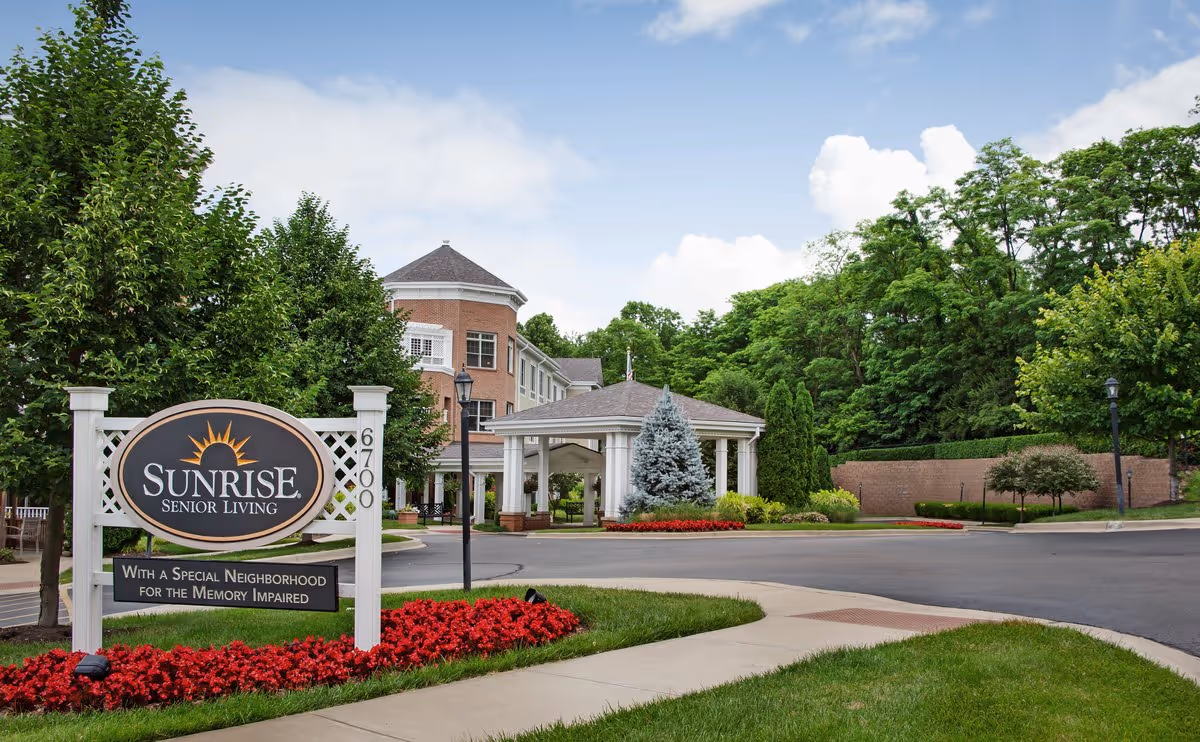 Exterior view of Sunrise Senior Living facility with a prominent sign in the foreground surrounded by red flowers and green trees. The building has a covered entrance and is set against a partly cloudy sky.