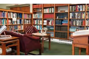 A cozy library room with wooden bookshelves filled with books lining the walls. There are two wooden tables with open books on them and a comfortable maroon armchair in the center. The room has a green carpet and a warm, inviting atmosphere.