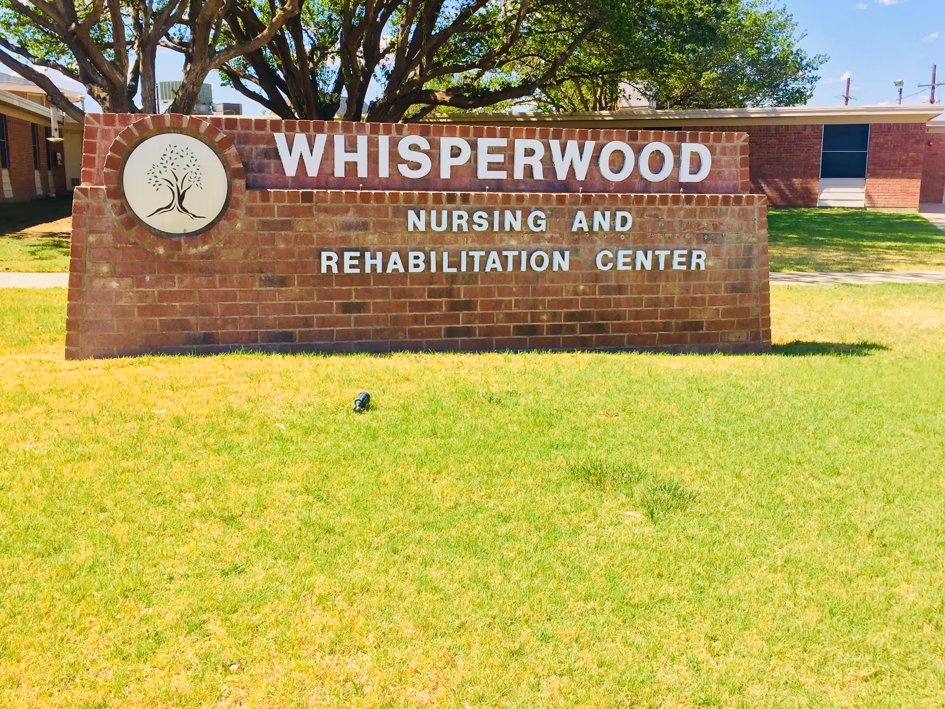 Brick sign on a grassy lawn that reads "Whisperwood Nursing and Rehabilitation Center" with a building and trees behind it.