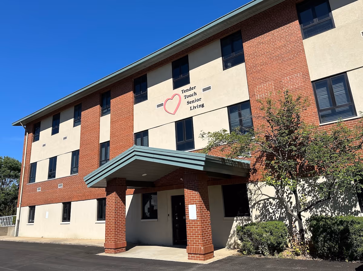 Exterior front of a three-story brick and stucco building with a covered entrance and a 'Tender Touch Senior Living' sign.