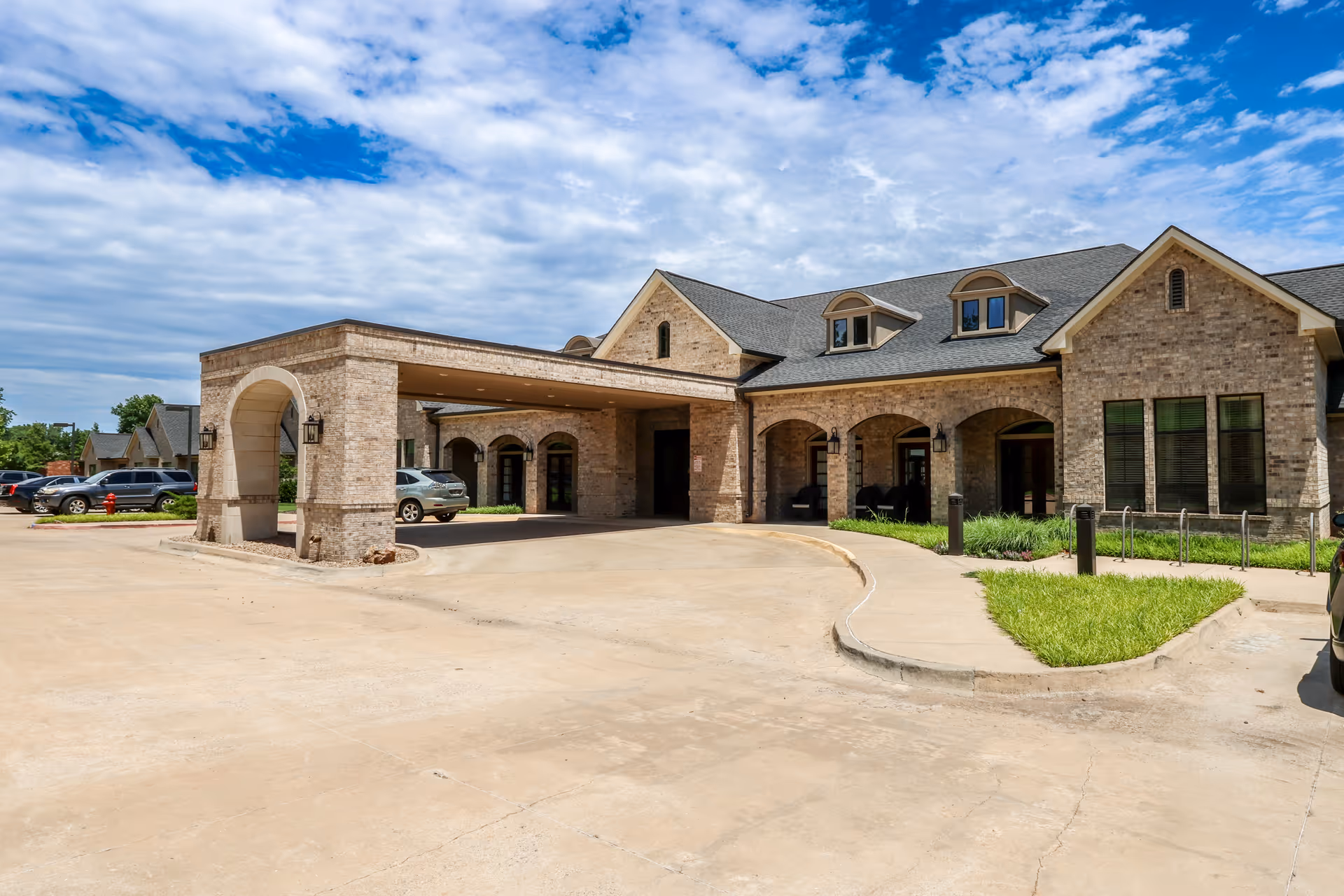 Exterior view of The Veraden Senior Living facility showing a large brick building with a covered entrance driveway, arched windows, and a well-maintained lawn under a partly cloudy sky.