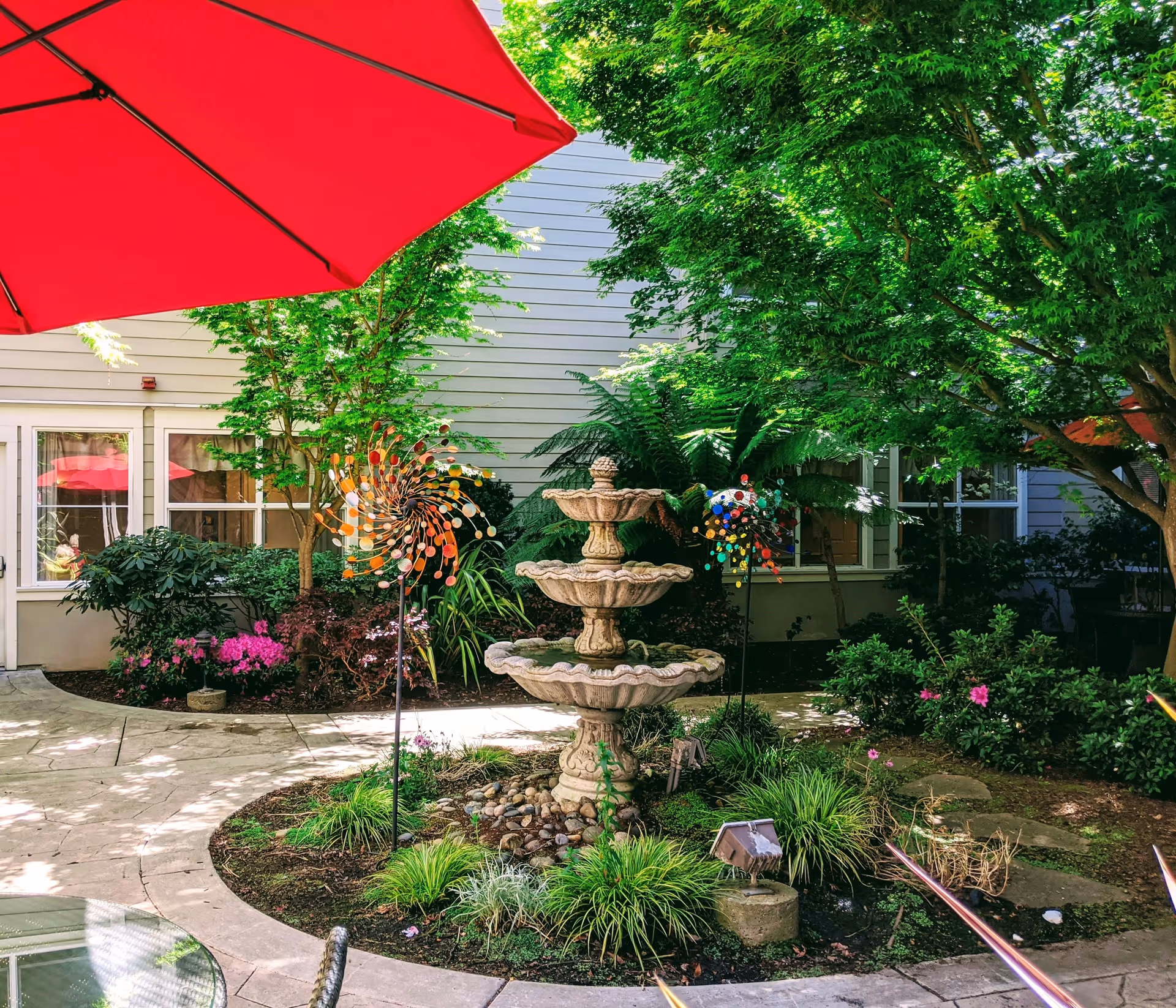 A peaceful outdoor courtyard featuring a three-tier stone fountain surrounded by green plants and colorful garden decorations. There is a large red umbrella providing shade, and the courtyard is bordered by a building with windows and doors.