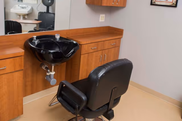 Salon-style hair washing station with a black shampoo bowl, wooden cabinets, mirror and a black barber chair.