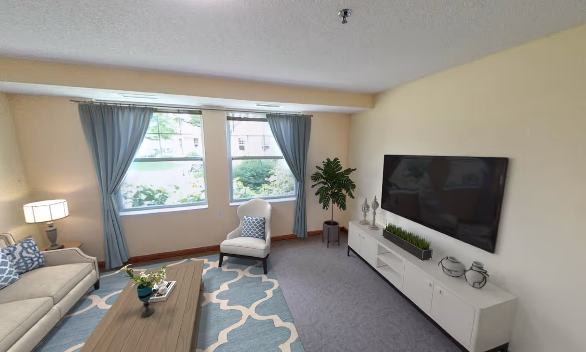 Well-lit living room with a sofa, armchair, coffee table, wall-mounted TV, and large windows with blue curtains.