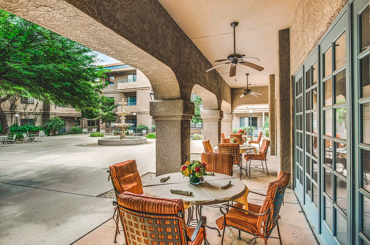 Outdoor covered patio area with round tables and cushioned chairs, each table decorated with a floral centerpiece. Ceiling fans hang from the patio roof. In the background, there is a courtyard with a multi-tiered water fountain, surrounded by trees and a building with balconies and windows.