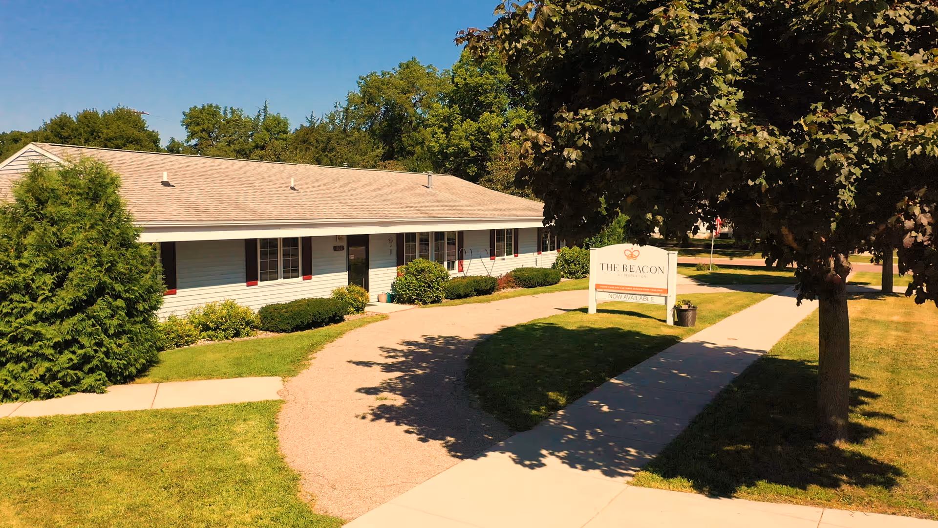 Exterior view of a single-story building with white siding and a gray roof, surrounded by green grass, bushes, and trees under a clear blue sky. A curved pathway leads to the entrance, and a sign near the sidewalk reads 'The Beacon at Mapleton' with additional text indicating availability.