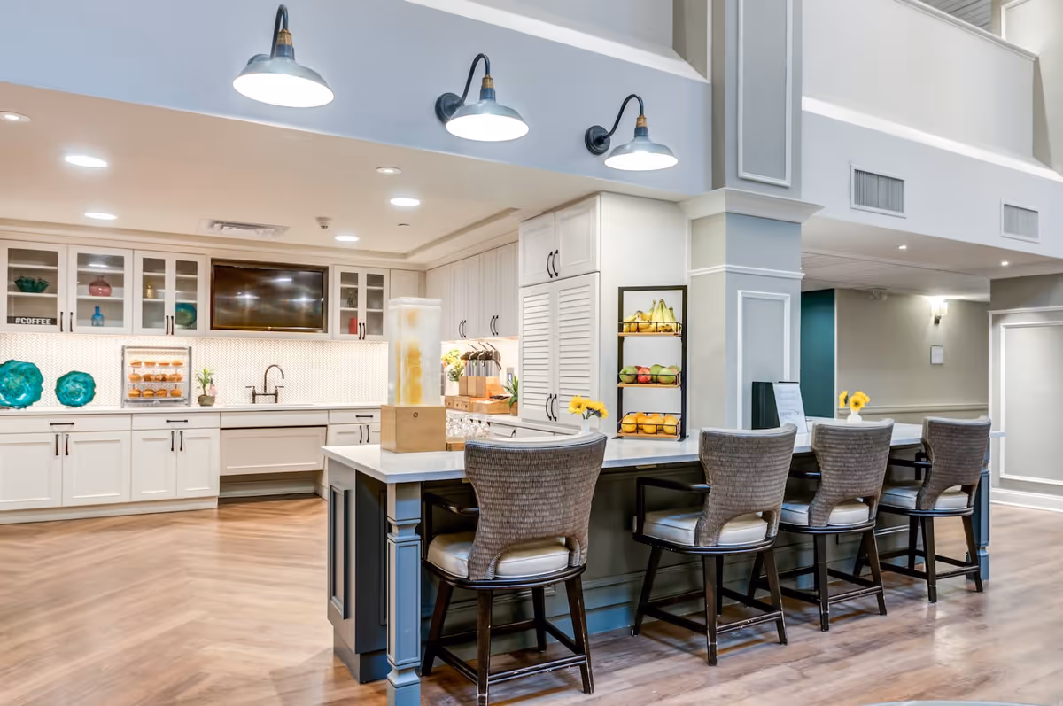 A modern kitchen area in a senior living facility with white cabinetry, a large island with four cushioned bar stools, pendant lights overhead, and a display of fresh fruit on a tiered stand. The space features wood flooring and a mounted flat-screen TV above the sink area.