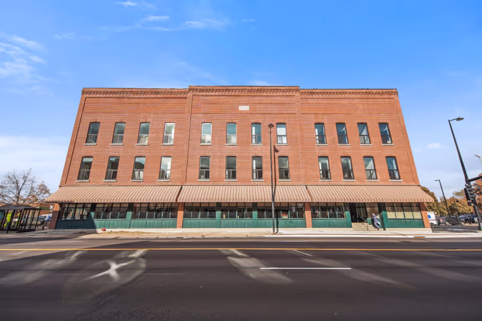Front exterior view of a three-story brick building with multiple windows and a striped awning covering the ground floor windows. A street with marked lanes runs in front of the building under a clear blue sky.