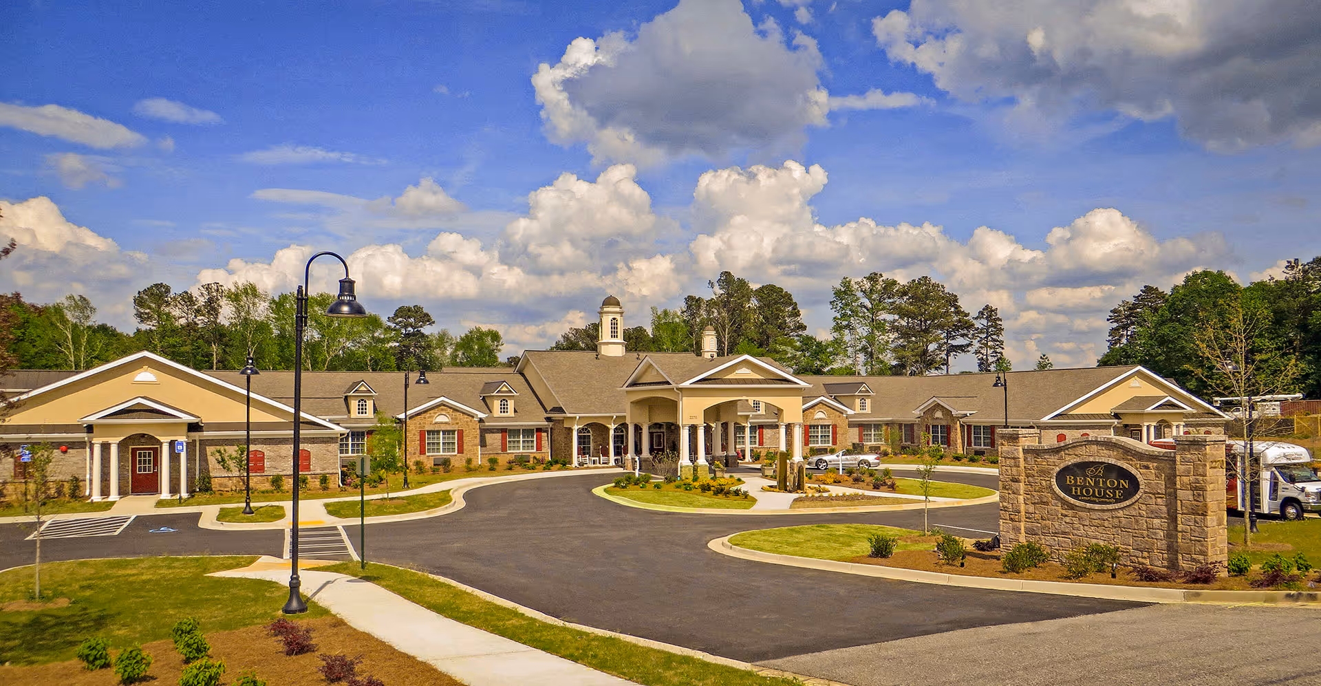 Exterior view of Benton House of Grayson, a single-story senior living facility with a large covered entrance, surrounded by landscaped grounds and a circular driveway under a partly cloudy blue sky.