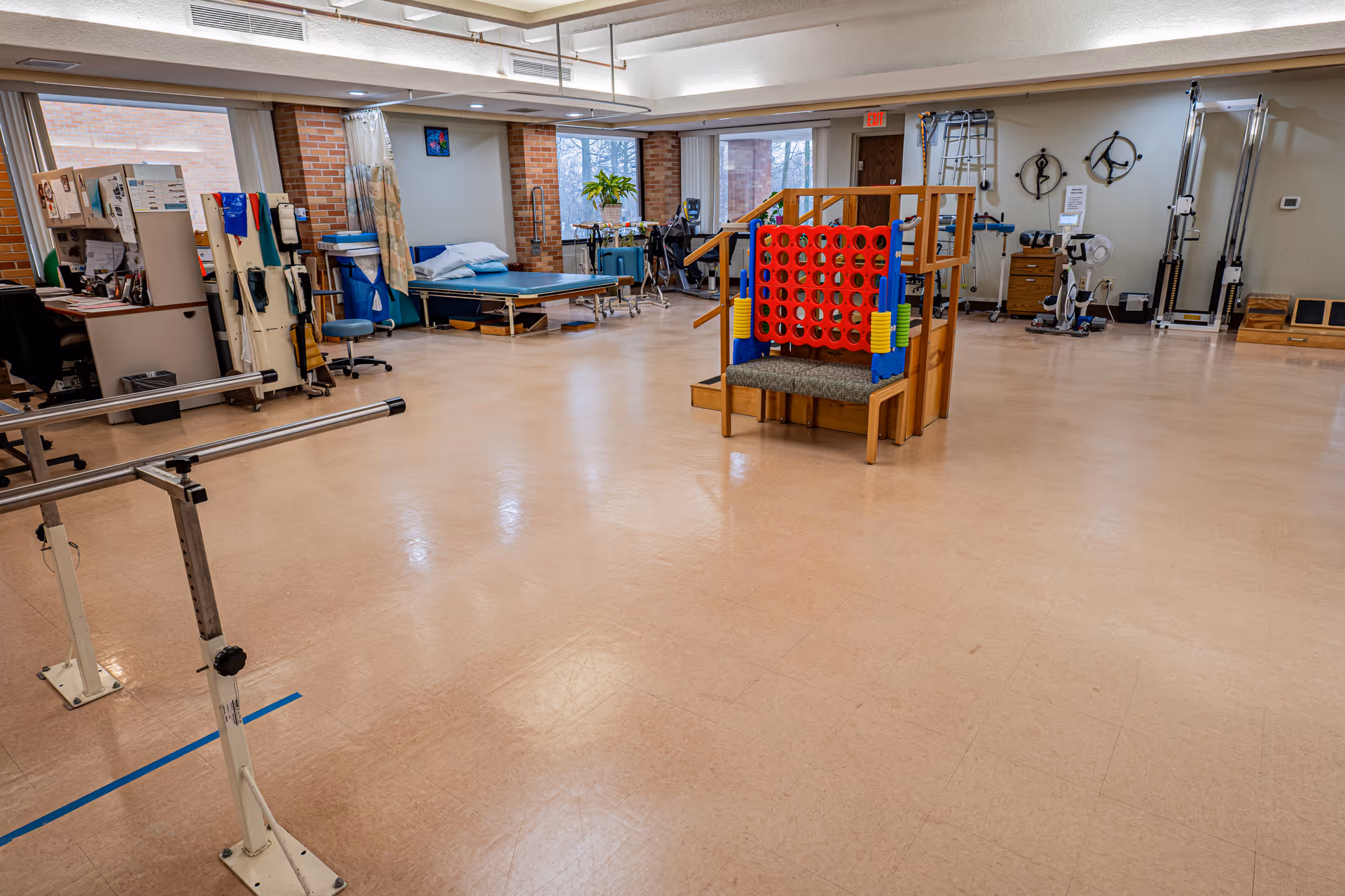 A spacious physical therapy room with exercise equipment including parallel bars, a large Connect Four game, therapy beds, and various rehabilitation tools. The room has large windows, brick walls, and a beige floor.
