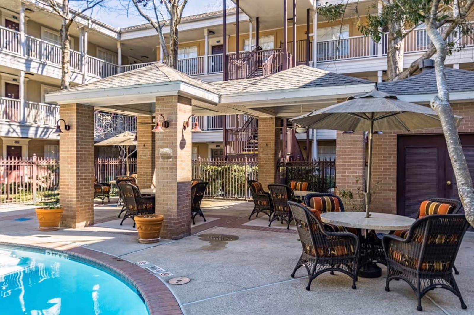 Outdoor seating area at King's Reserve Willowbrook 55+ Active Adult Living featuring wicker chairs with cushions around tables, some shaded by umbrellas, next to a swimming pool and surrounded by a multi-story building with balconies.