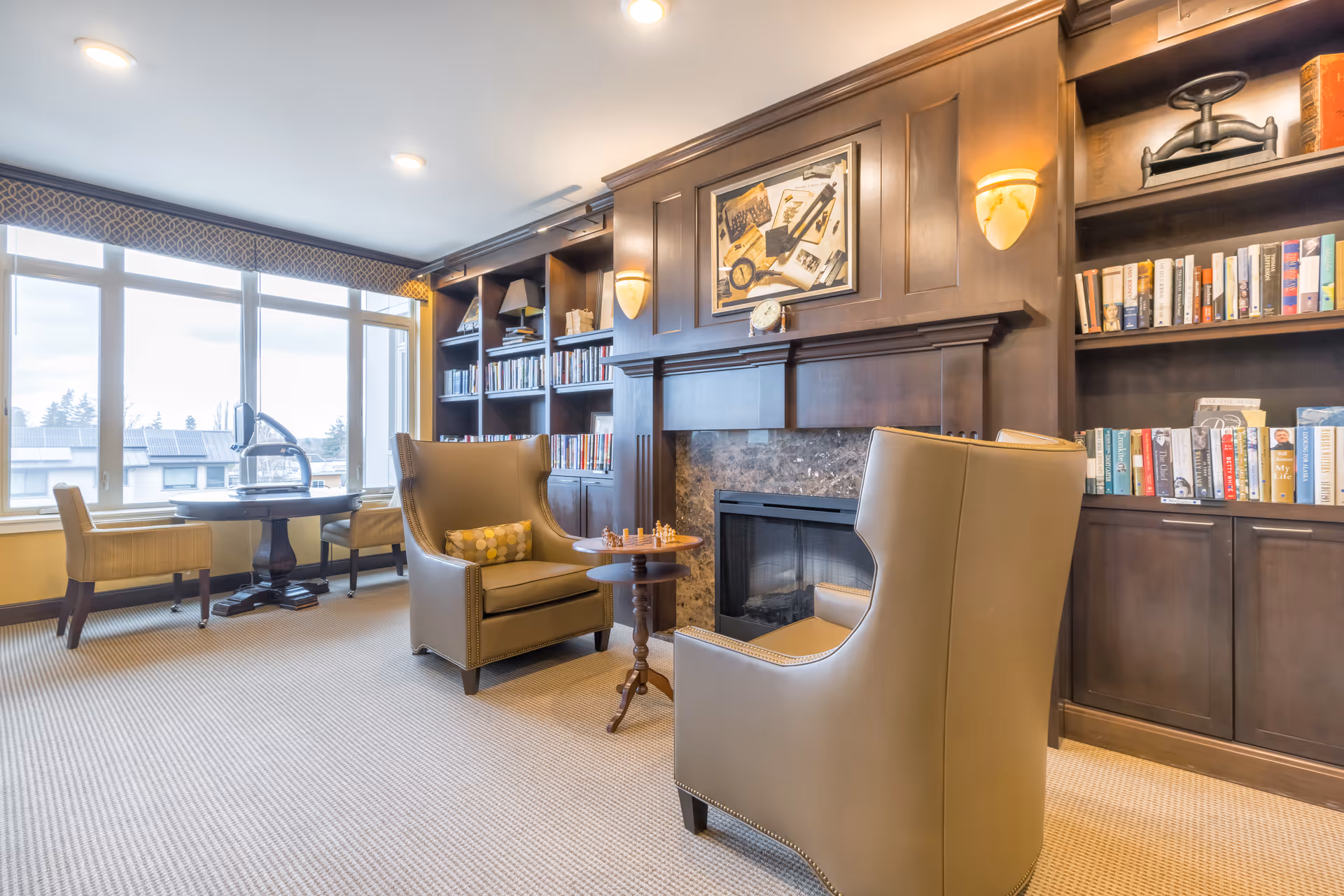 A cozy living room area with two beige armchairs facing a small round table with a chess set. Behind the chairs is a fireplace with a dark wooden mantel and built-in bookshelves filled with books and decorative items. Large windows on the left side allow natural light to fill the room.
