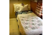 A small single bed with a quilted patchwork blanket, pillows, and a curtained window in a modest bedroom.