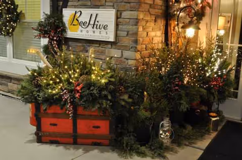 Festive exterior entry with illuminated potted greenery and BeeHive Homes sign on brick wall