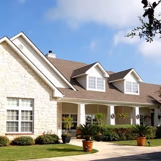 Front exterior of a single-story residential building with a covered porch, dormer windows, potted plants, and a manicured lawn.
