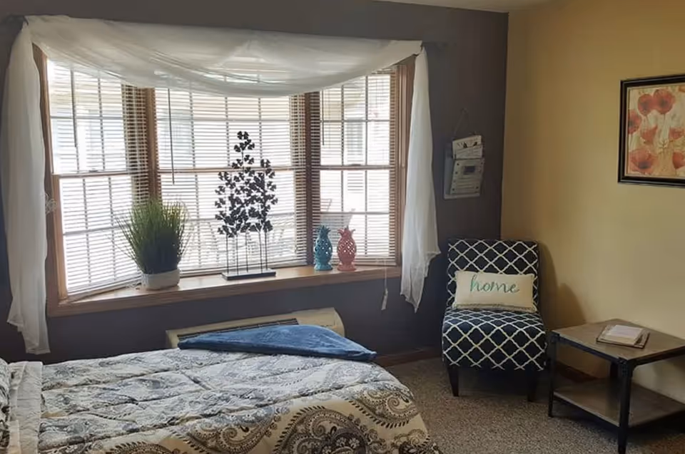 A cozy bedroom with a large bay window featuring wooden blinds and white sheer curtains. A bed with a patterned comforter is partially visible in the foreground. On the window sill, there are decorative items including a potted plant and two pineapple-shaped ornaments. To the right, there is a patterned armchair with a pillow that says 'home' and a small side table with a book on it. The walls are painted in two tones, dark brown and beige, and a framed floral picture hangs on the beige wall.