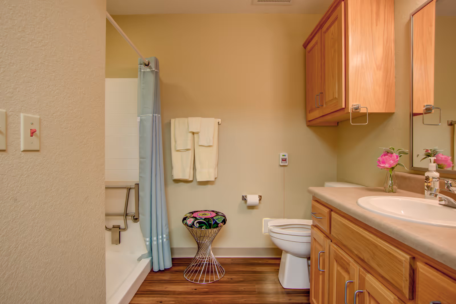 A bathroom with a wooden vanity and sink on the right, a toilet next to it, a colorful stool in front of the toilet, and a bathtub with a blue shower curtain on the left. Two white towels hang on a towel rack above the stool, and a small vase with pink flowers is placed on the vanity countertop.