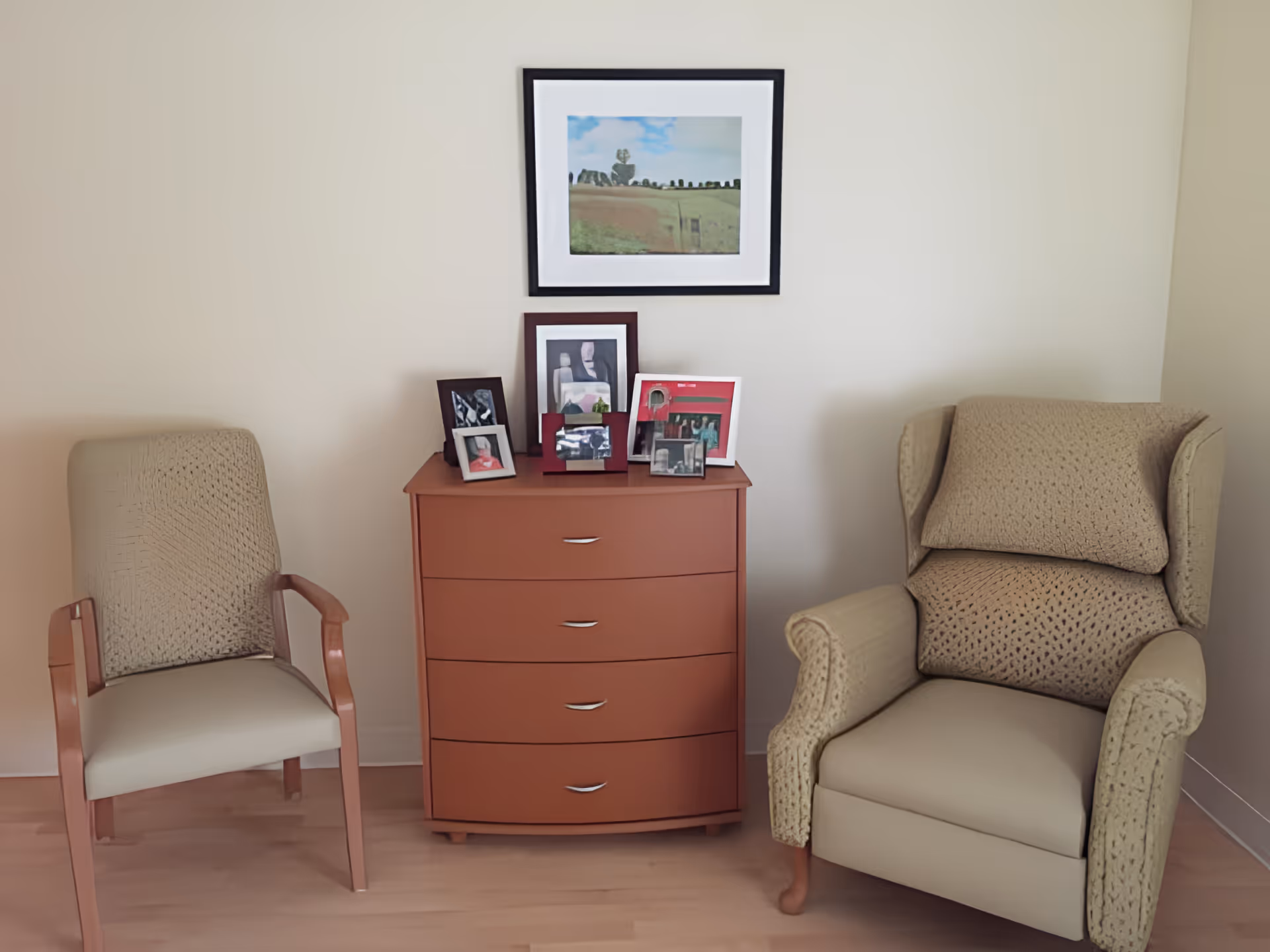 Two upholstered chairs flank a wooden dresser topped with framed photos and a landscape picture hanging above on a neutral wall.