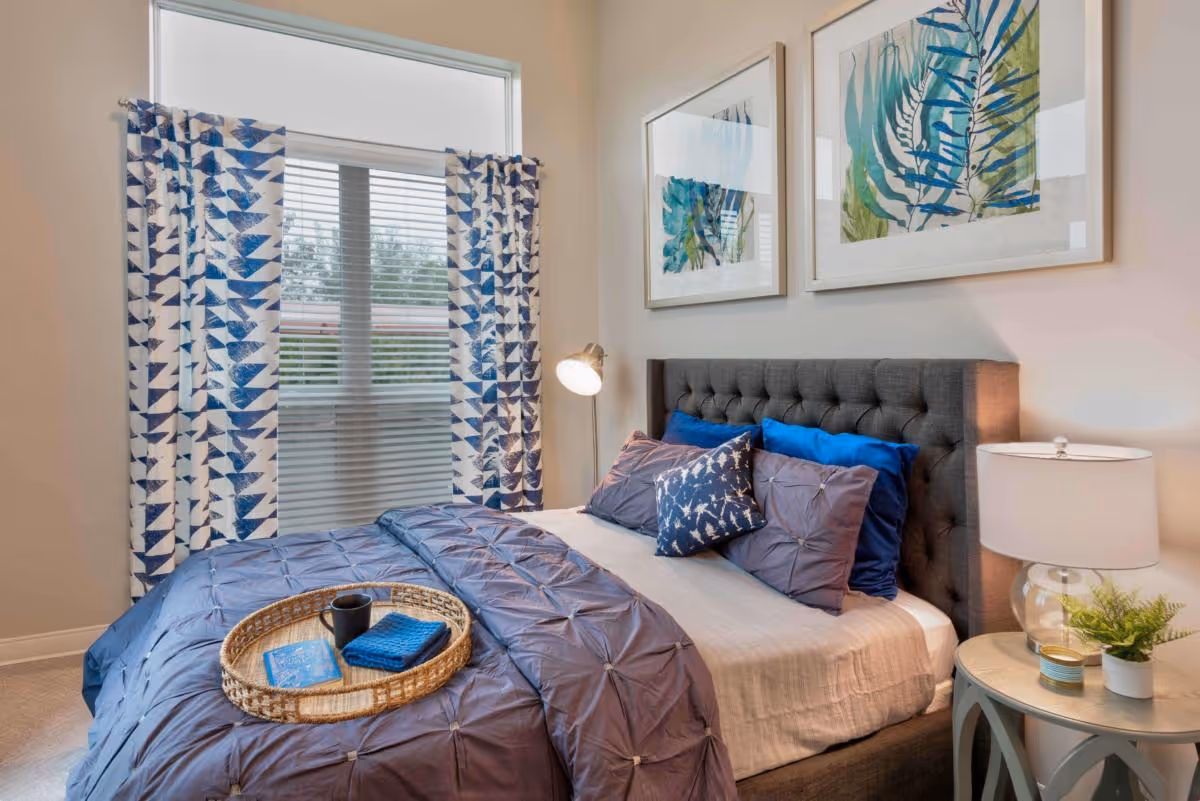 A cozy bedroom with a neatly made bed featuring blue and gray bedding and decorative pillows. A wicker tray with a book, a cup, and a cloth is placed on the bed. The room has a window with blue and white patterned curtains, two framed botanical prints on the wall above the bed, a floor lamp, and a round bedside table with a lamp, a small plant, and a candle.