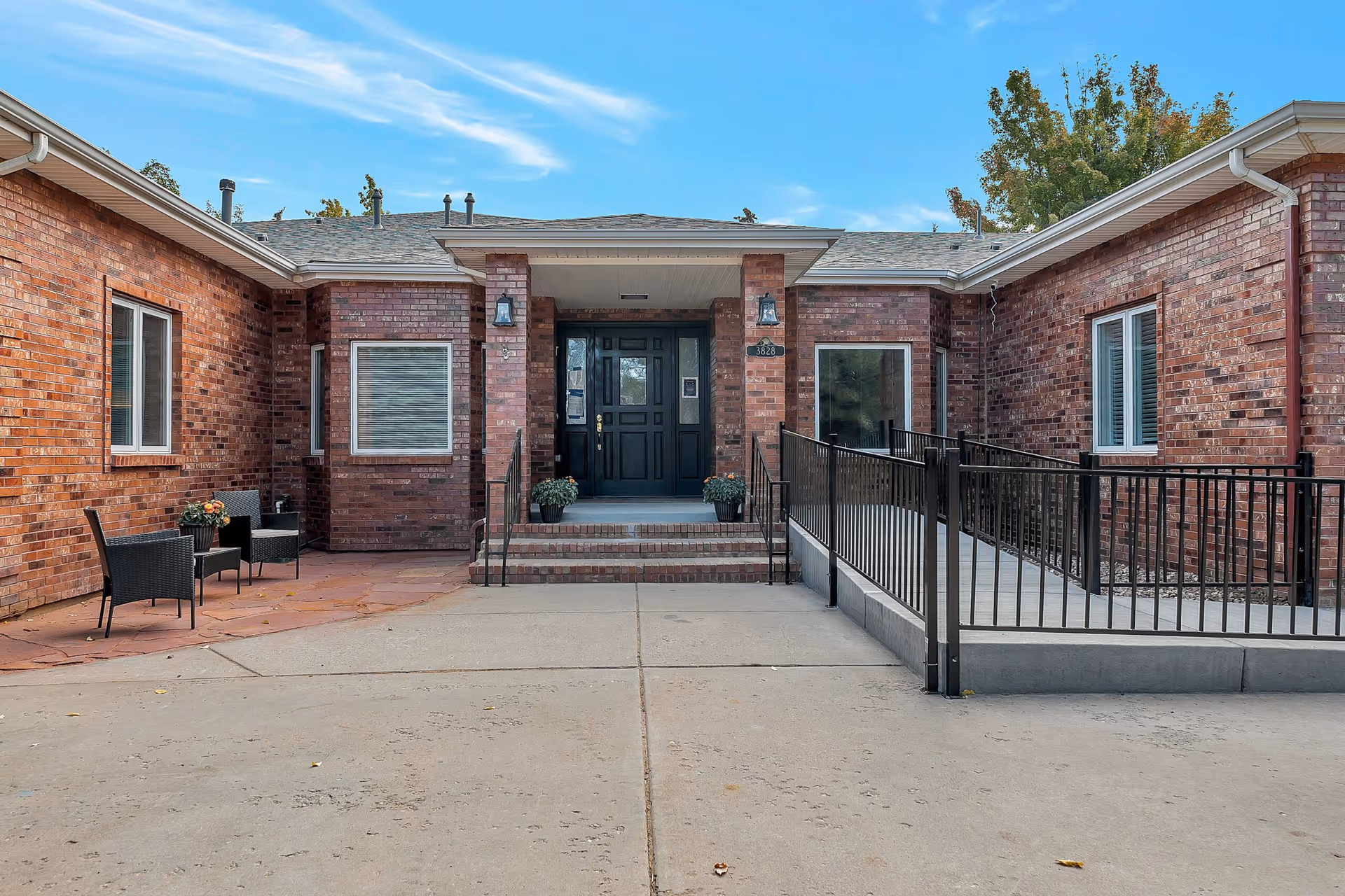 Front entrance of a brick building with a black door, two potted plants on the steps, a wheelchair accessible ramp with black railings on the right, and two black chairs with a small table on the left side under windows. The sky is clear with some clouds and there are trees in the background.