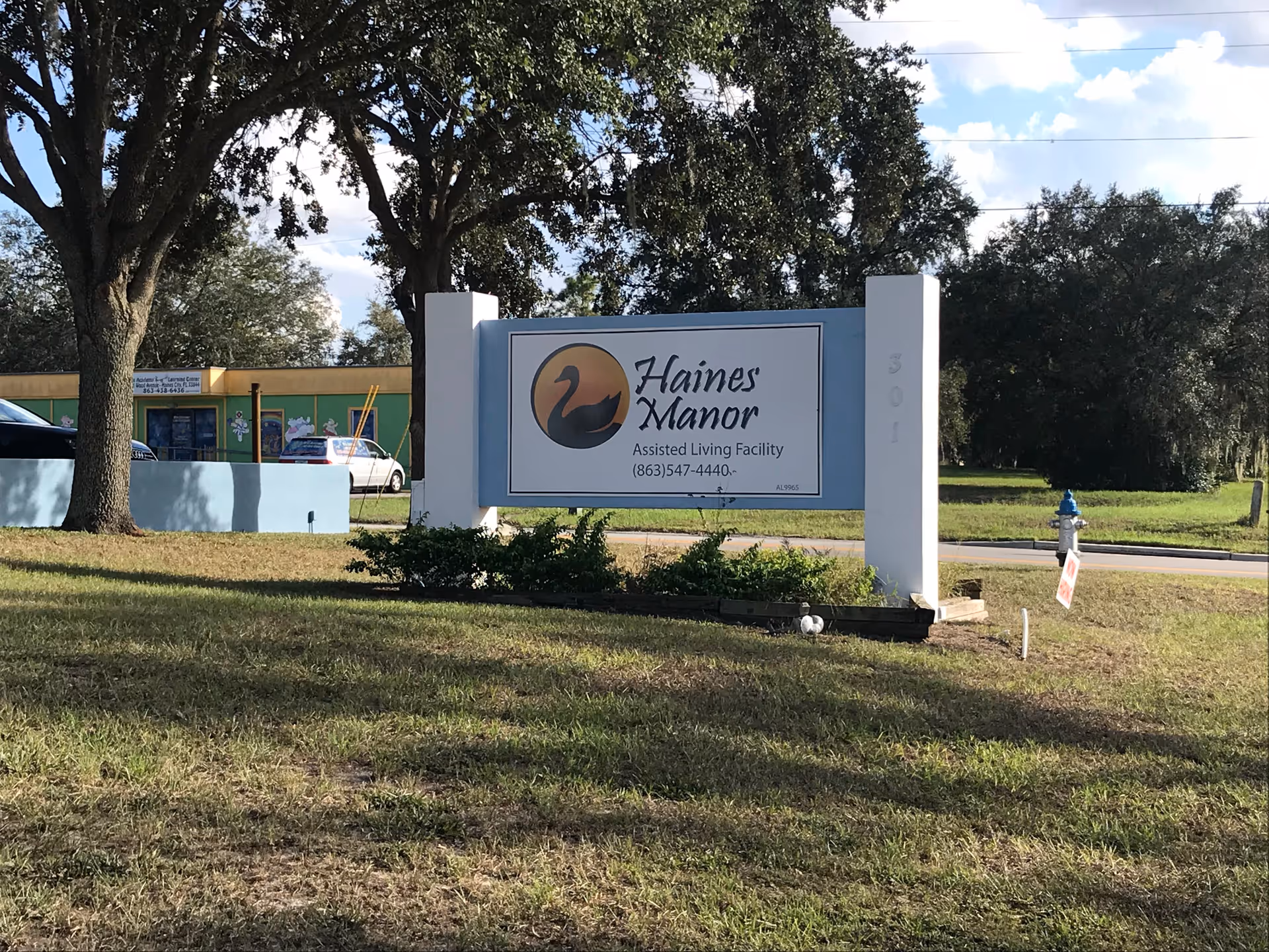 Roadside sign for Haines Manor Assisted Living on a grassy lawn with trees and a building in the background.