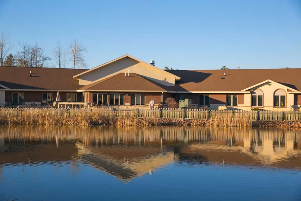 Exterior view of a single-story senior living facility building with a brown roof and brick walls, reflected in a calm pond in the foreground. There is a wooden fence and some tall grasses along the edge of the pond, with clear blue sky above.