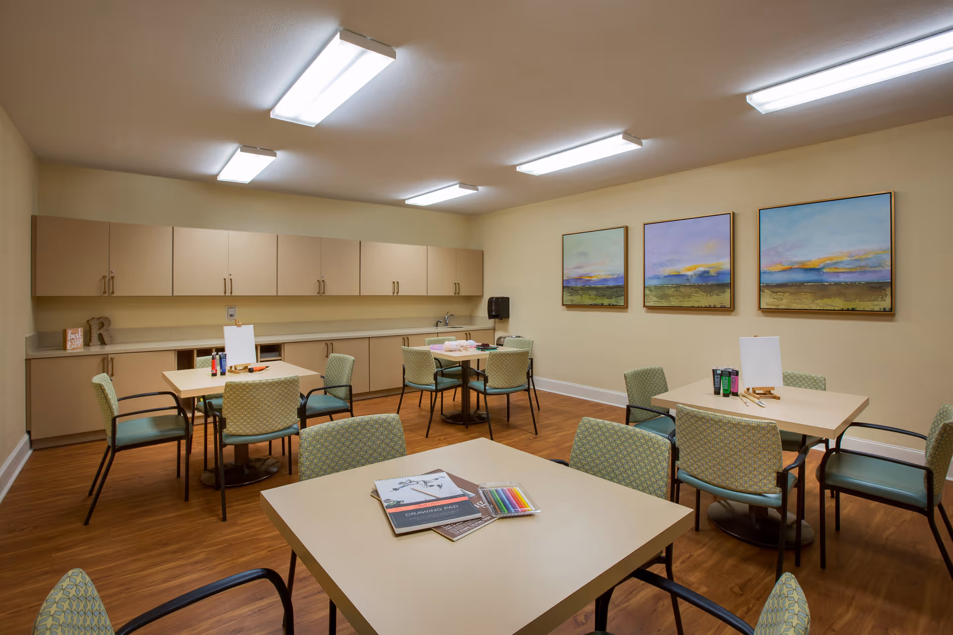 A well-lit activity room with several tables and chairs arranged for group activities. The tables have art supplies like colored pencils, paint tubes, and blank canvases. The room has beige walls, wooden flooring, and a long countertop with cabinets above and below along one wall. Three landscape paintings hang on one wall.