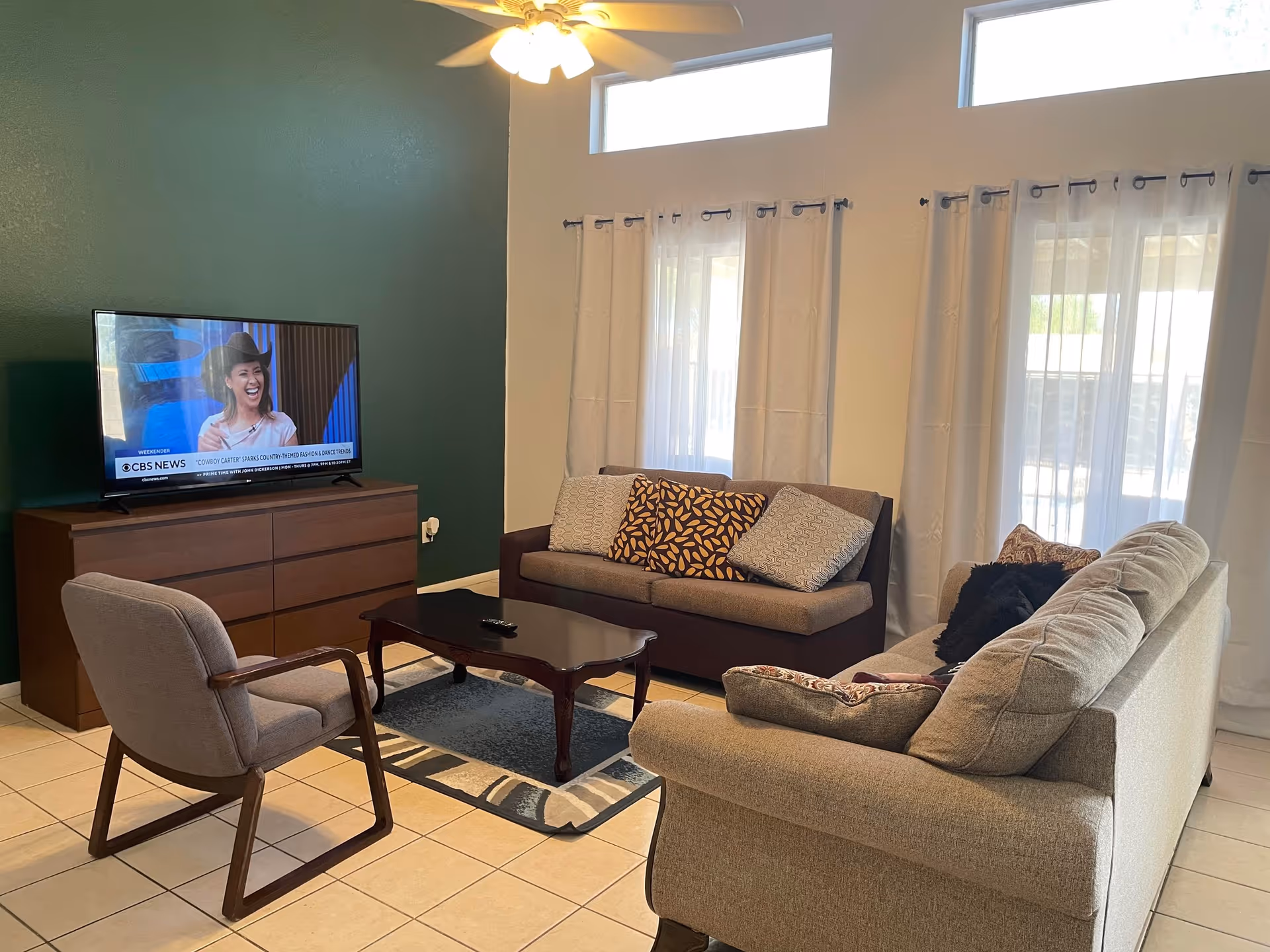 A cozy living room with a beige sofa, a dark brown loveseat with patterned cushions, a gray armchair, and a wooden coffee table on a rug. A flat-screen TV on a wooden dresser is showing a CBS News segment. The room has large windows with sheer white curtains and a ceiling fan with lights.