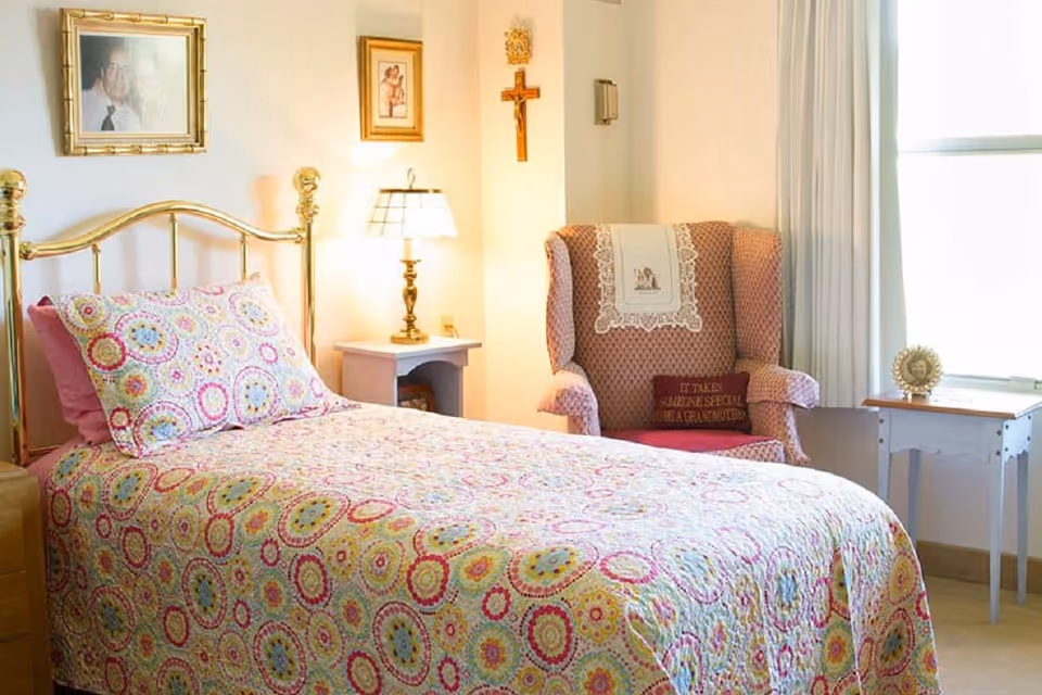 Bright bedroom featuring a brass-framed bed with a colorful patterned quilt, a nightstand with a lamp, and an upholstered armchair by a window.