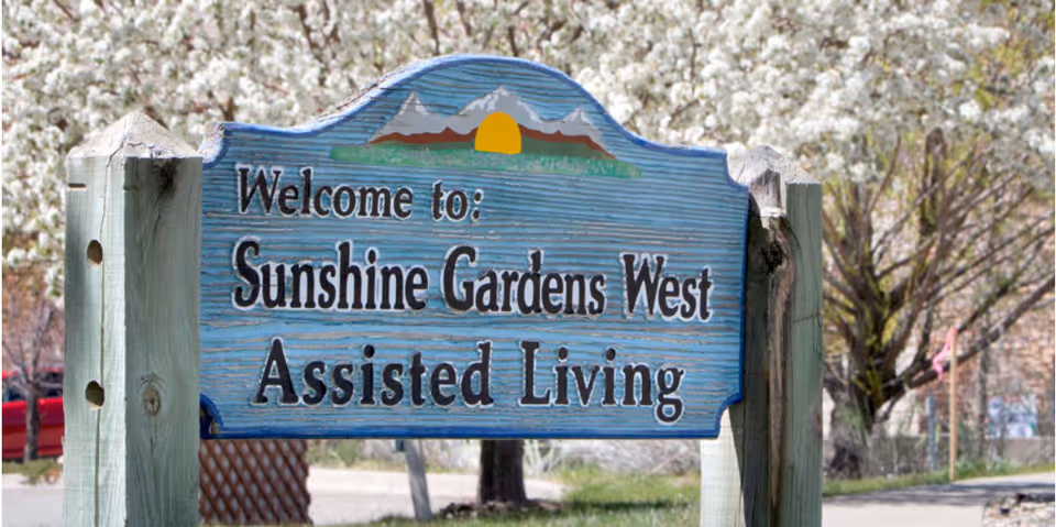 A wooden sign painted blue with a sun and mountain design at the top, reading 'Welcome to: Sunshine Gardens West Assisted Living', set outdoors with trees and a road in the background.