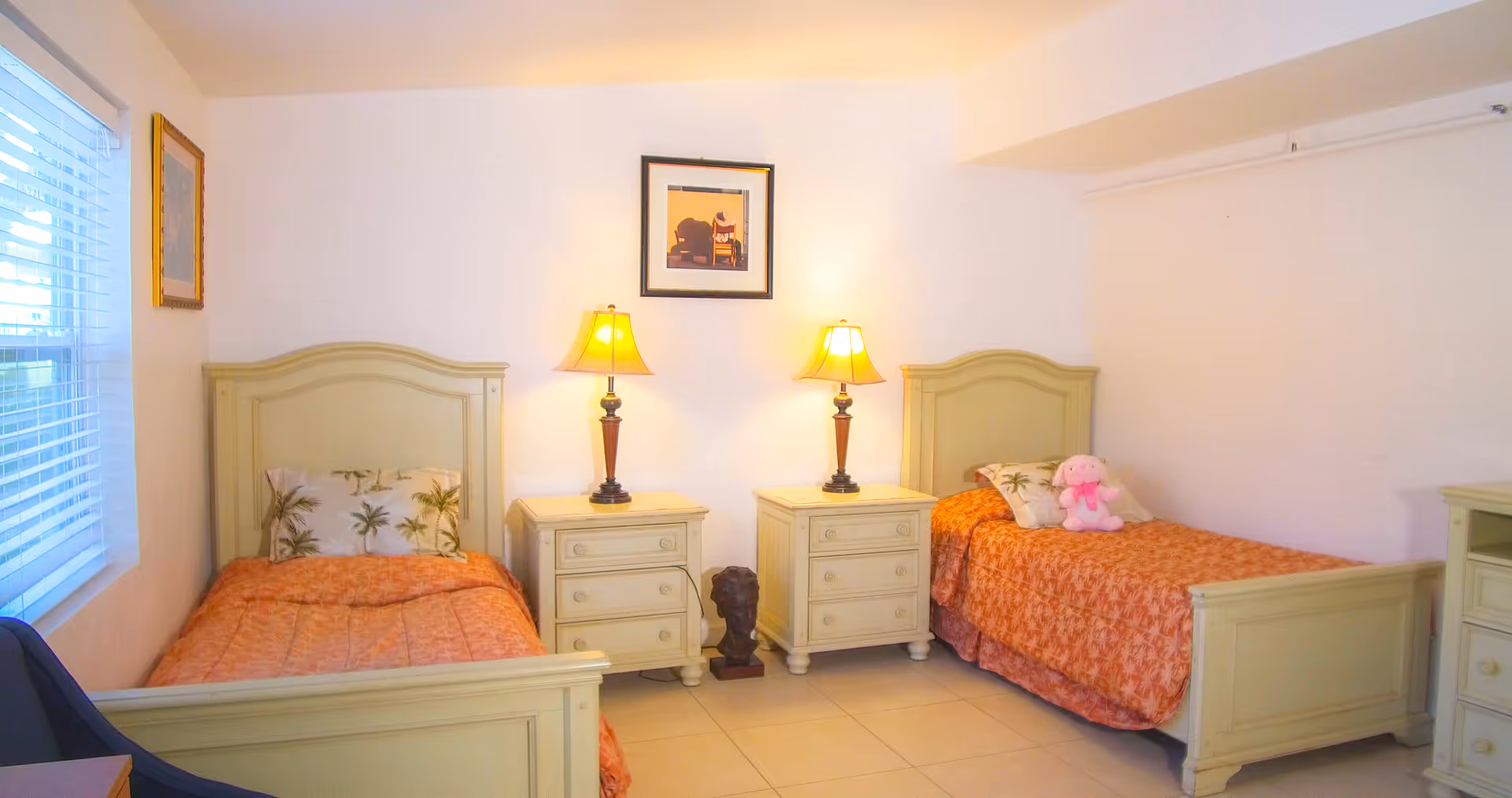 A bedroom with two single beds, each with a light green wooden frame and orange bedspreads. There are two matching nightstands between the beds, each with a lamp on top. A framed picture hangs on the wall above the nightstands. A window with blinds is on the left side of the room, and a pink stuffed animal is placed on the right bed.