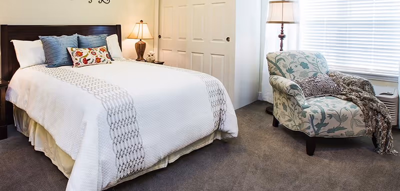 A cozy bedroom featuring a neatly made bed with white and patterned bedding, several pillows, a bedside table with a lamp, a floral patterned armchair with a throw blanket, a floor lamp, and a window with blinds.